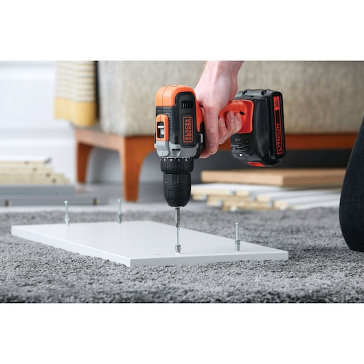 BLACKDECKER BCD703C1 cordless drill driving a screw into white wood panel on carpeted floor.