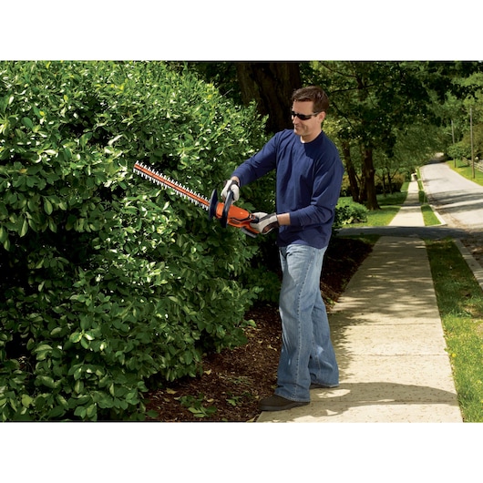 BLACKDECKER BCHT222D1 hedge trimmer used outdoors, cutting green bushes along a sidewalk.
