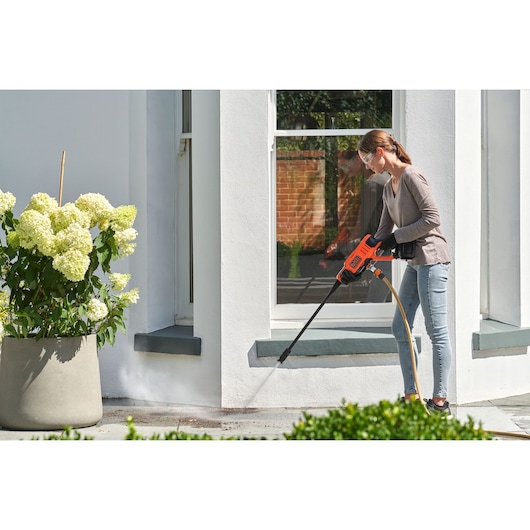Photo of a person cleaning a patio with a BLACKDECKER BCPW350E1 pressure washer near a white wall and potted plant.