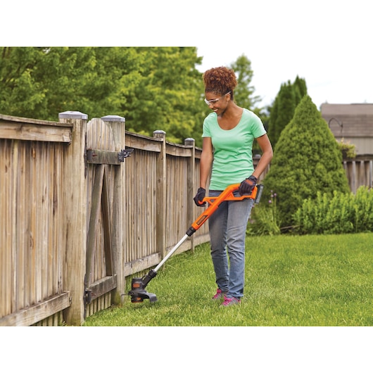 Photo of person using BLACKDECKER BCST201C1 orange cordless string trimmer on lawn near wooden fence.