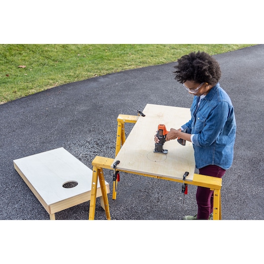 BLACKDECKER BDCDMT1206KITC power tool used for cutting a circular hole in a wood board on sawhorses outdoors.