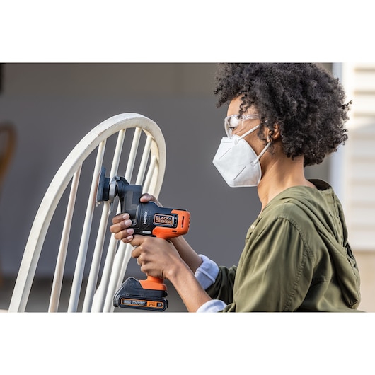 Person sanding a wooden chair with BLACK+DECKER BDCDMT1206KITC cordless power tool, wearing safety goggles and mask.