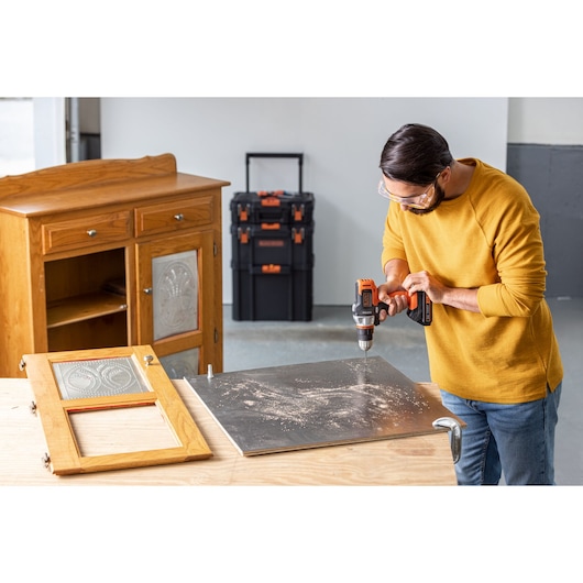 Photo of person using BLACKDECKER BDCDMT1206KITC cordless drill on metal sheet with wood cabinet and toolboxes in background.