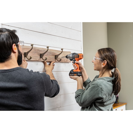 Photo of a woman using a BLACK+DECKER BDCDMT1206KITC cordless drill to mount a wooden coat rack on a white wall.