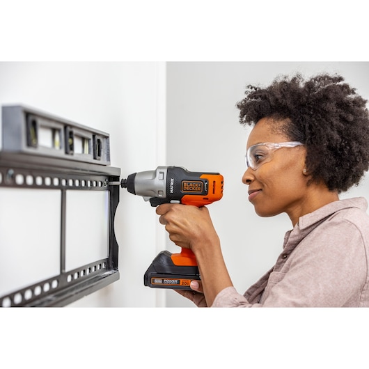 Photo of a person using a BLACK+DECKER BDCDMT1206KITC cordless drill to mount a TV bracket on a white wall.