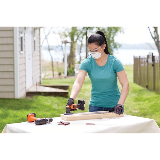 Photo of person using BLACKDECKER BDCK502C1 cordless sander on wood outdoors, drill and sandpaper on table.