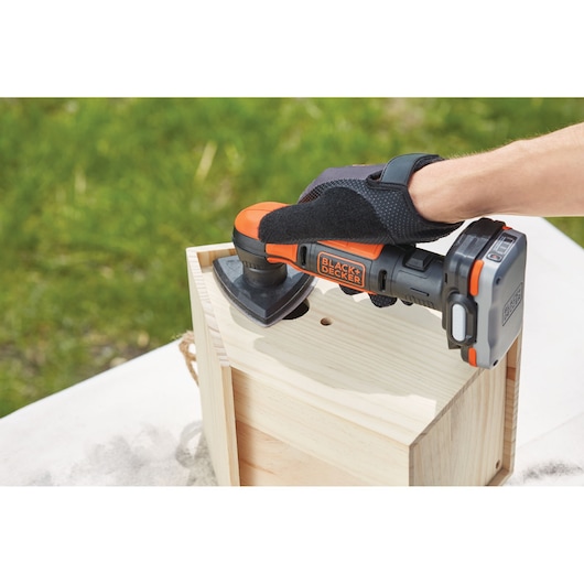 Photo of BLACK+DECKER BDCK502C1 cordless detail sander in use on wooden box, outdoor angle.