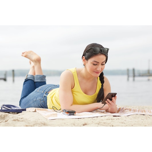 BLACKDECKER BDCK502C1 portable charger powering a smartphone, used by a woman lying on a beach towel by the water.