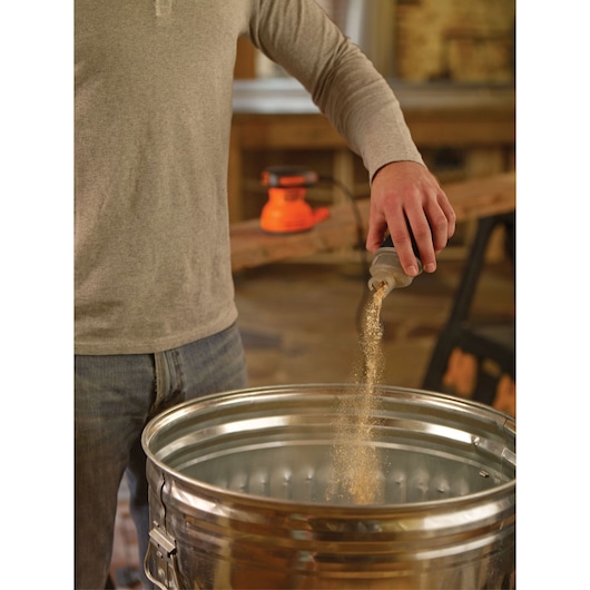 Photo of person emptying dust from BLACKDECKER BDERO100 sander container into a metal trash can in workshop.