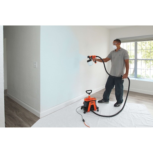 Photo of BLACKDECKER BDPH1200 paint sprayer being used to paint an indoor wall, unit on the floor with hose connected.