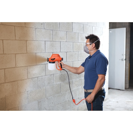 BLACKDECKER BDPS130 handheld paint sprayer in use painting a concrete wall, orange and black design.