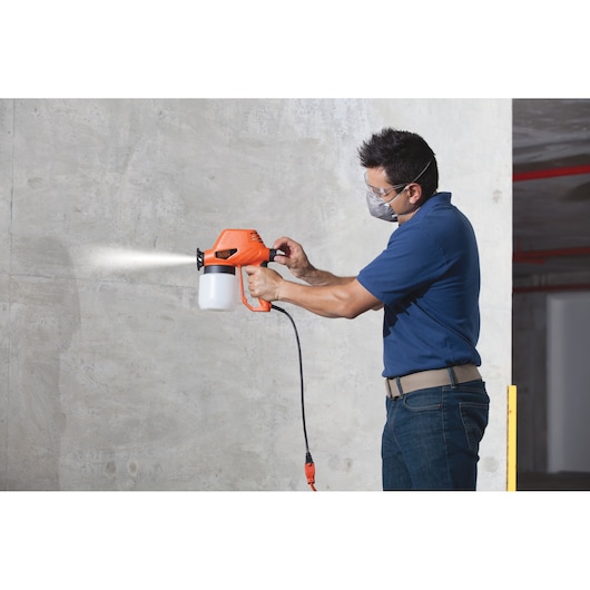Person using BLACKDECKER BDPS130 electric paint sprayer, orange and black, spraying on a concrete wall.