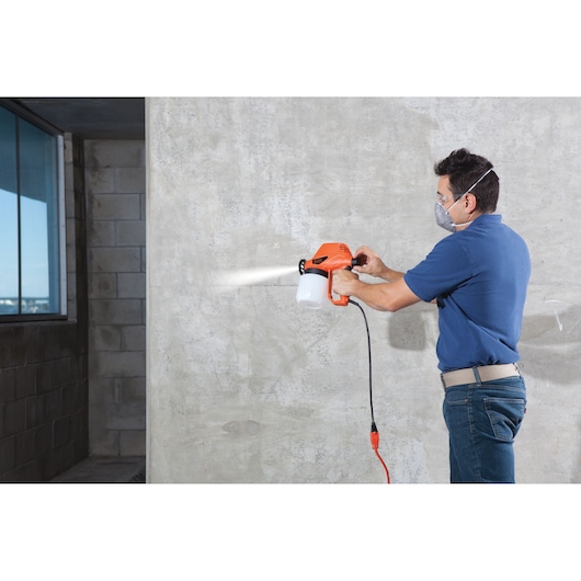 Photo of a person using BLACKDECKER BDPS130 paint sprayer with orange body and white canister on a concrete wall.
