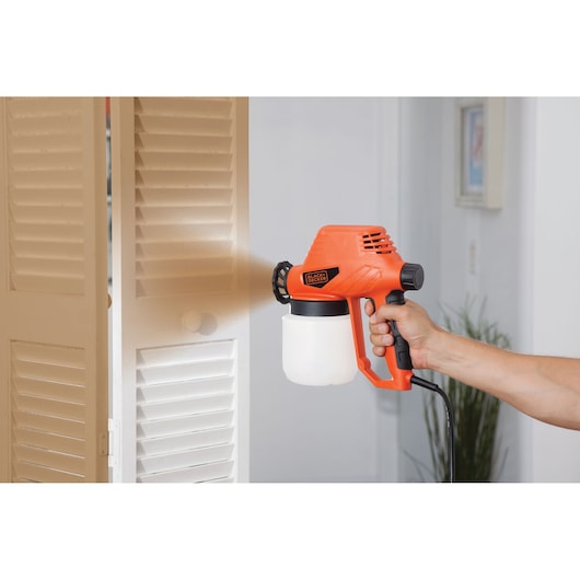 BLACKDECKER BDPS130 handheld paint sprayer in use painting a white louvered door indoors.