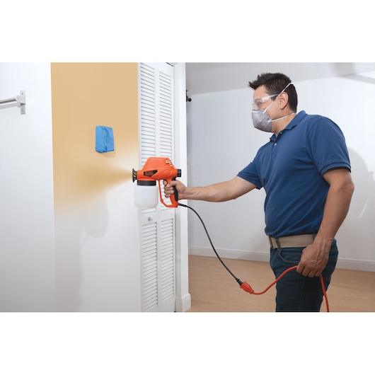 Person using BLACKDECKER BDPS130 paint sprayer to apply beige paint on indoor wall, holding orange tool with mask on.