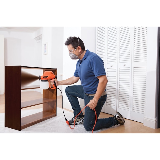 Person using BLACKDECKER BDPS130 paint sprayer to finish a wooden shelf, interior setting, side angle.