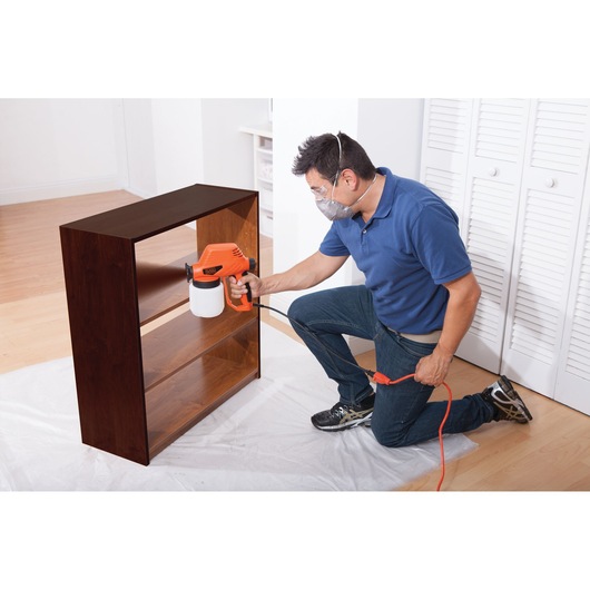 Photo of person using BLACKDECKER BDPS130 paint sprayer on wooden shelf in indoor setting.