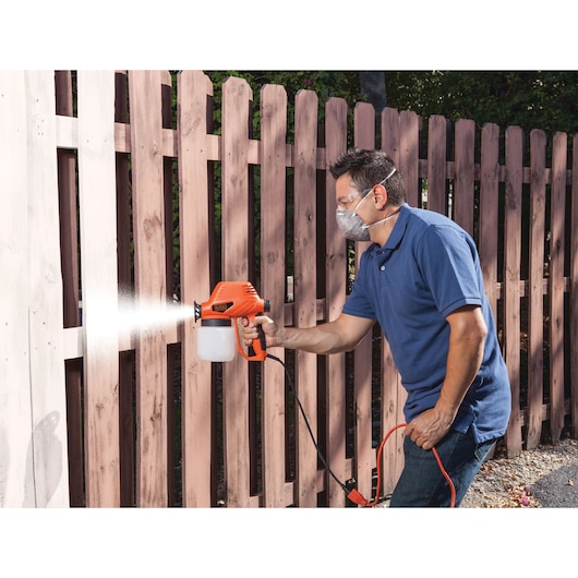 BLACKDECKER BDPS130 paint sprayer in use painting a wooden fence outdoors, user wearing a blue shirt.
