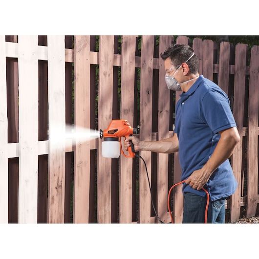 Photo of person using BLACKDECKER BDPS130 paint sprayer to paint a wooden fence outdoors.