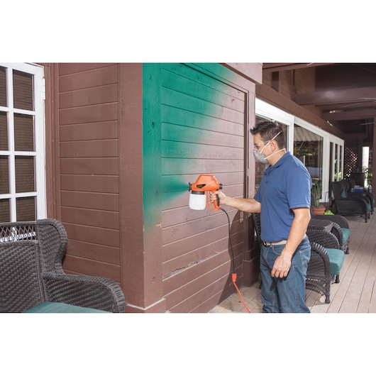 Person using BLACKDECKER BDPS130 paint sprayer to apply green paint to brown outdoor wooden wall.