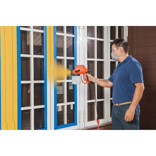 Photo of a person using a BLACKDECKER BDPS130 paint sprayer on a yellow-trim window, spraying yellow paint outdoors.