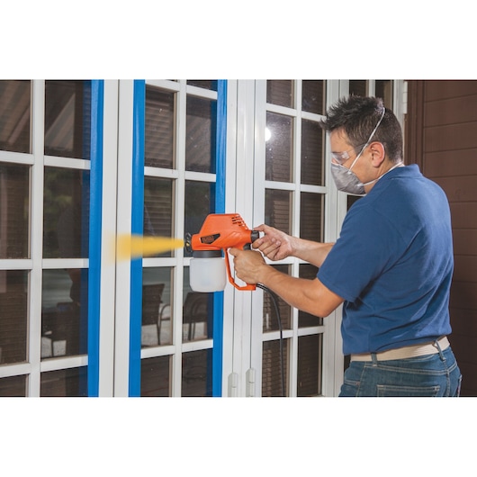 Photo of person using BLACKDECKER BDPS130 paint sprayer on white framed glass doors with blue masking tape.