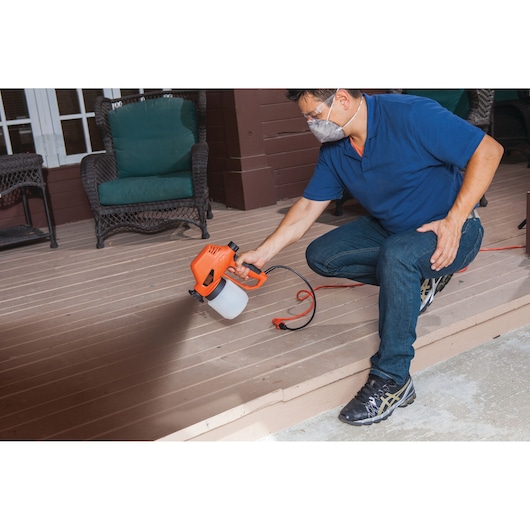 Person using BLACKDECKER BDPS130 paint sprayer on outdoor deck, orange tool with white paint container.