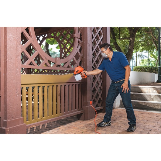 Photo of a person using the BLACKDECKER BDPS130 electric paint sprayer to paint a wooden fence outdoors.
