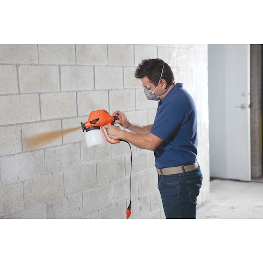 Person using BLACKDECKER BDPS130 paint sprayer on concrete wall indoors.