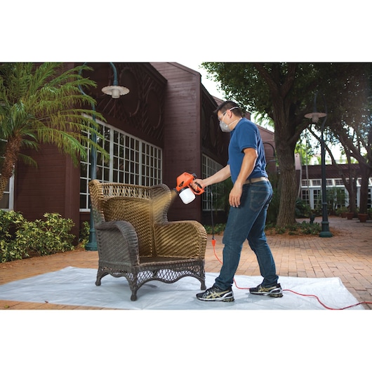 Photo of person using BLACKDECKER BDPS130 paint sprayer to refinish outdoor wicker chair on patio.