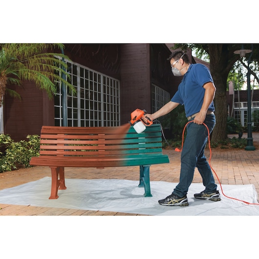 BLACKDECKER BDPS130 paint sprayer used to paint a wooden bench outdoors, orange and black tool with cord.