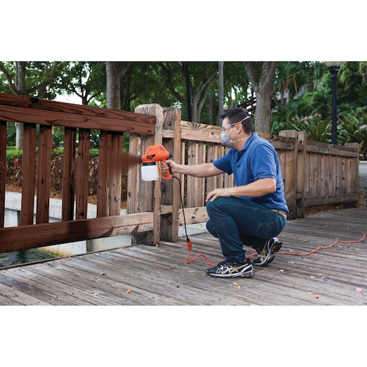 Photo of person using BLACKDECKER BDPS130 paint sprayer to stain wooden fence outdoors.