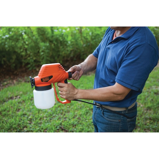 Photo of a person holding a BLACK+DECKER BDPS130 orange electric paint sprayer outdoors, with a grass background.