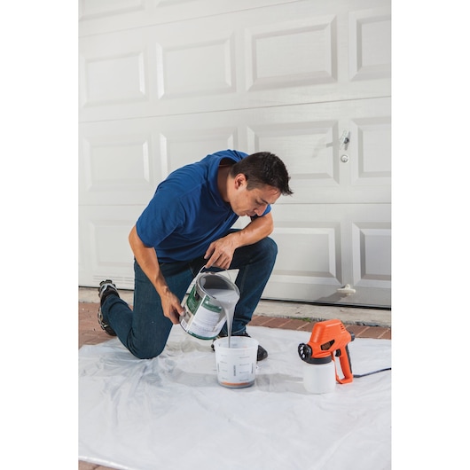 Photo of person pouring paint into a container beside a BLACKDECKER paint sprayer BDPS130 in front of a garage door.