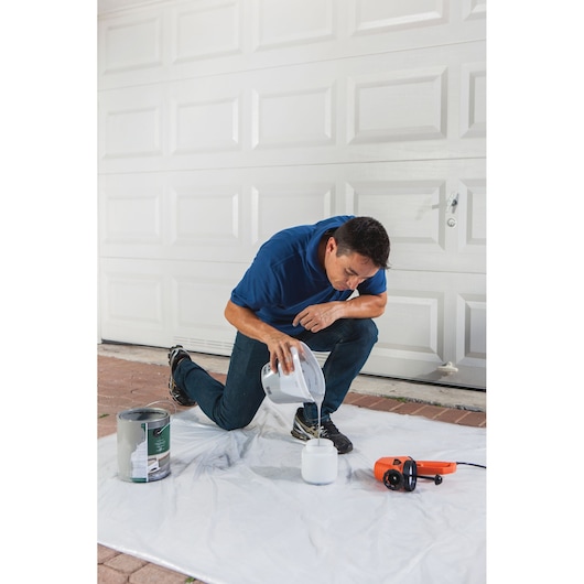 Photo of person filling a paint container next to BLACKDECKER BDPS130 paint sprayer and paint can, outdoor setup.