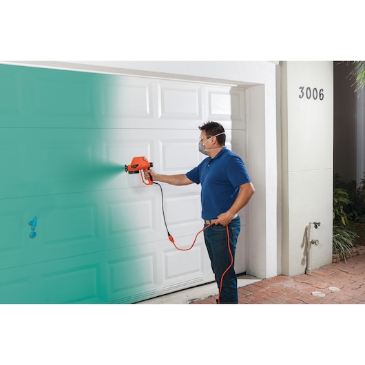 BLACKDECKER BDPS130 paint sprayer being used to spray blue paint on a white garage door outdoors.