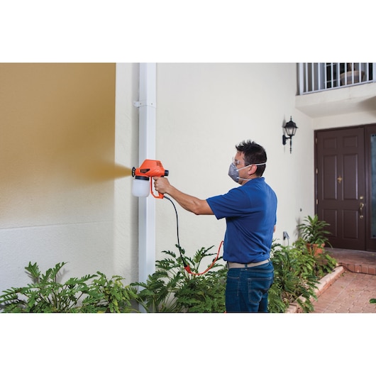 BLACKDECKER BDPS130 handheld paint sprayer in use painting exterior wall, orange and white design.
