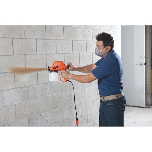 Photo of a person using BLACKDECKER BDPS130 paint sprayer on a concrete wall indoors.