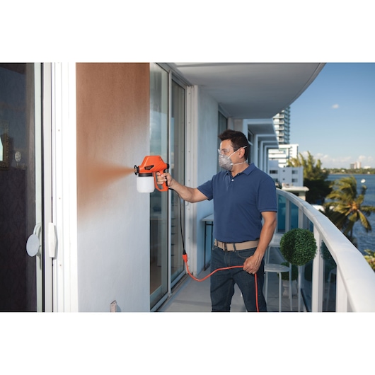 Photo of person using BLACKDECKER BDPS130 paint sprayer to spray paint a white exterior wall on a balcony.