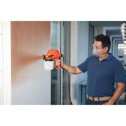 Photo of BLACKDECKER BDPS130 paint sprayer in use, spraying paint on a white wall outdoors.