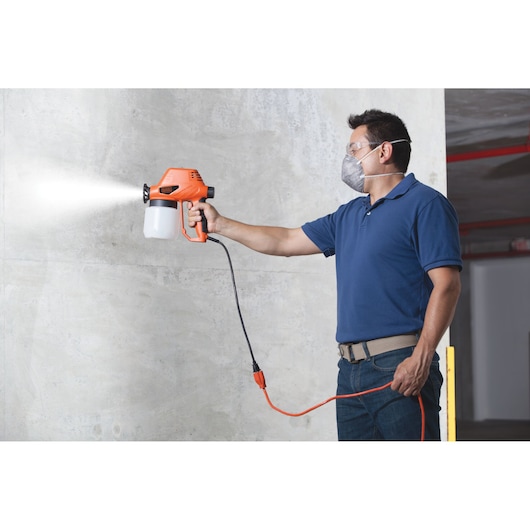 Person using BLACKDECKER BDPS130 electric paint sprayer with orange body and white container on indoor wall.
