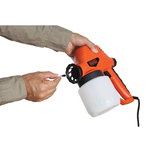 Photo of hands adjusting BLACKDECKER BDPS130 paint sprayer with orange body and white paint container, side angle.