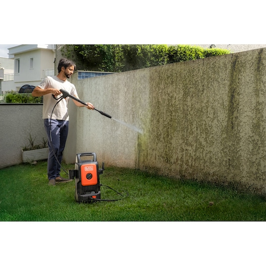 Photo of a person using BLACK+DECKER BEPW1300 pressure washer cleaning an exterior wall in a garden.