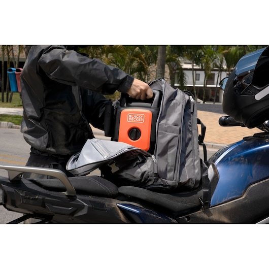 BLACK+DECKER BEPW1300 portable pressure washer being placed in a gray backpack on a motorcycle seat outdoors.