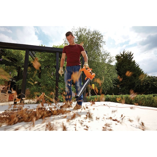 Photo of person using BLACKDECKER BV25 leaf blower on patio, orange and black tool blowing dry leaves outdoors.