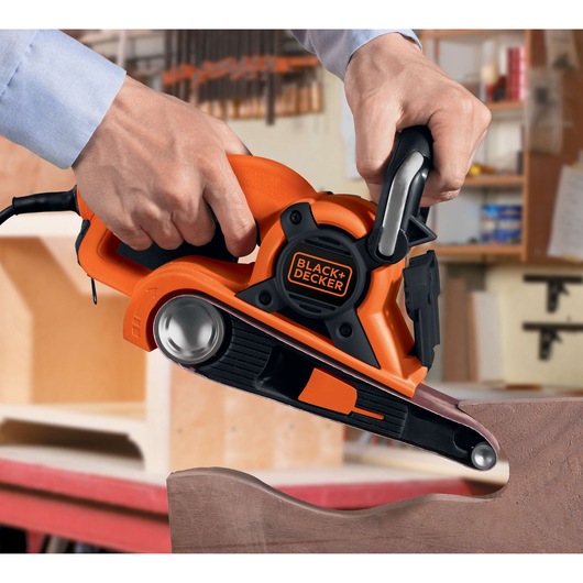 BLACK+DECKER DS321 belt sander in use, orange and black design, sanding curved wooden edge in workshop.