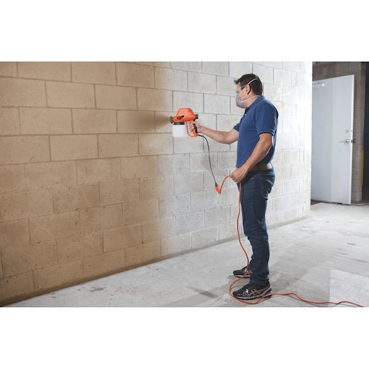 Photo of person using BLACKDECKER BDPS130 paint sprayer on concrete wall indoors.