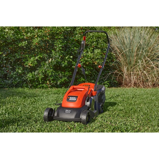 A BLACK+DECKER electric lawn mower with a bright orange and black design is shown on a grassy lawn with green shrubs in the background.