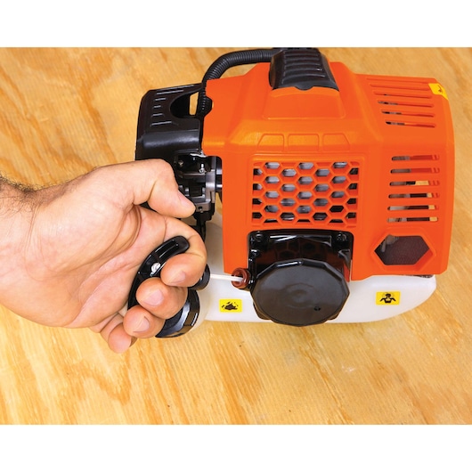 Photo of BLACKDECKER GST33 brush cutter engine close-up on wood surface, hand pulling starter cord, orange and black housing.
