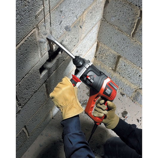 Photo of BLACK+DECKER KD975KA rotary hammer used to chisel a hole in a gray concrete block wall, operator wearing gloves.